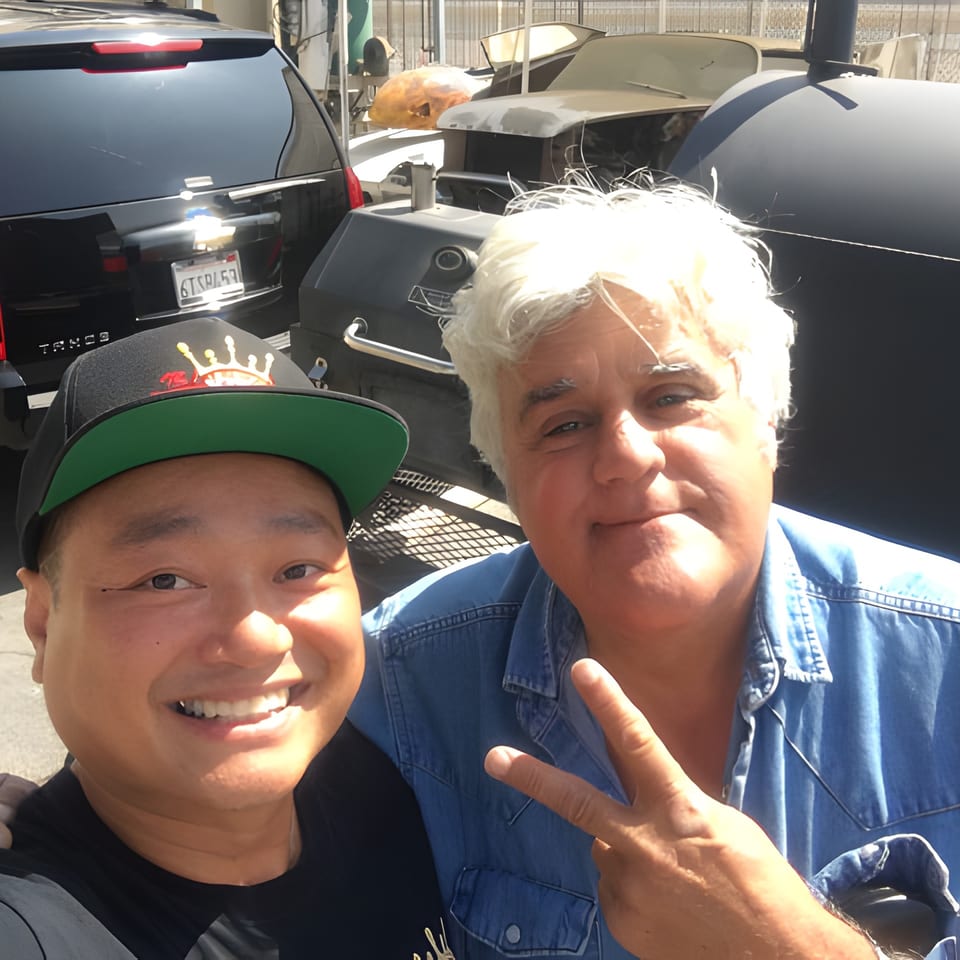 Jay Leno and a man smiling for a selfie with cars and large BBQ smokers.