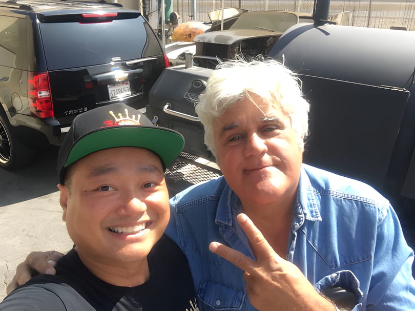Jay Leno and a man smiling for a selfie with cars and large BBQ smokers.