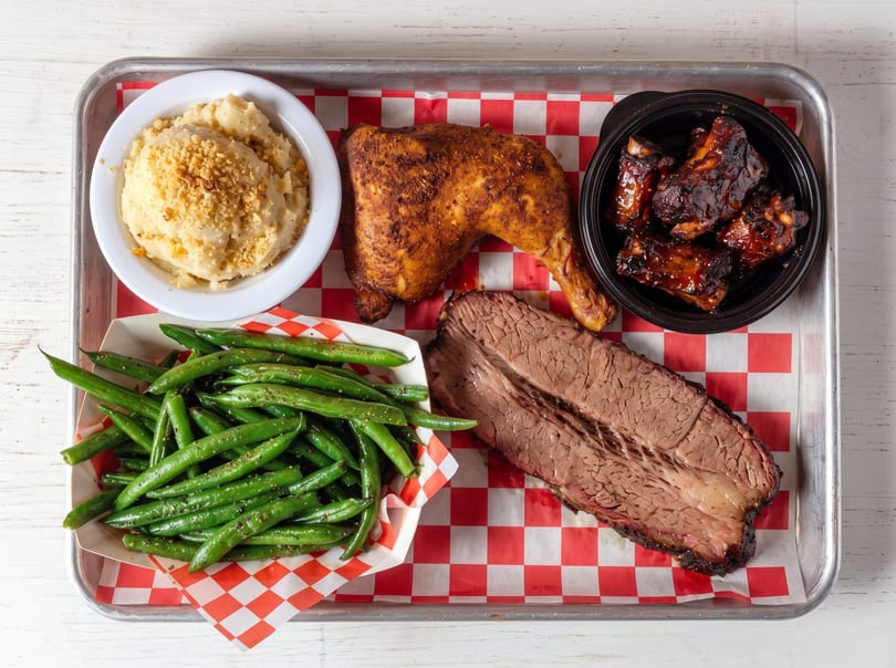 Barbecue platter with chicken, ribs, brisket, mashed potatoes, and green beans on a metal tray.