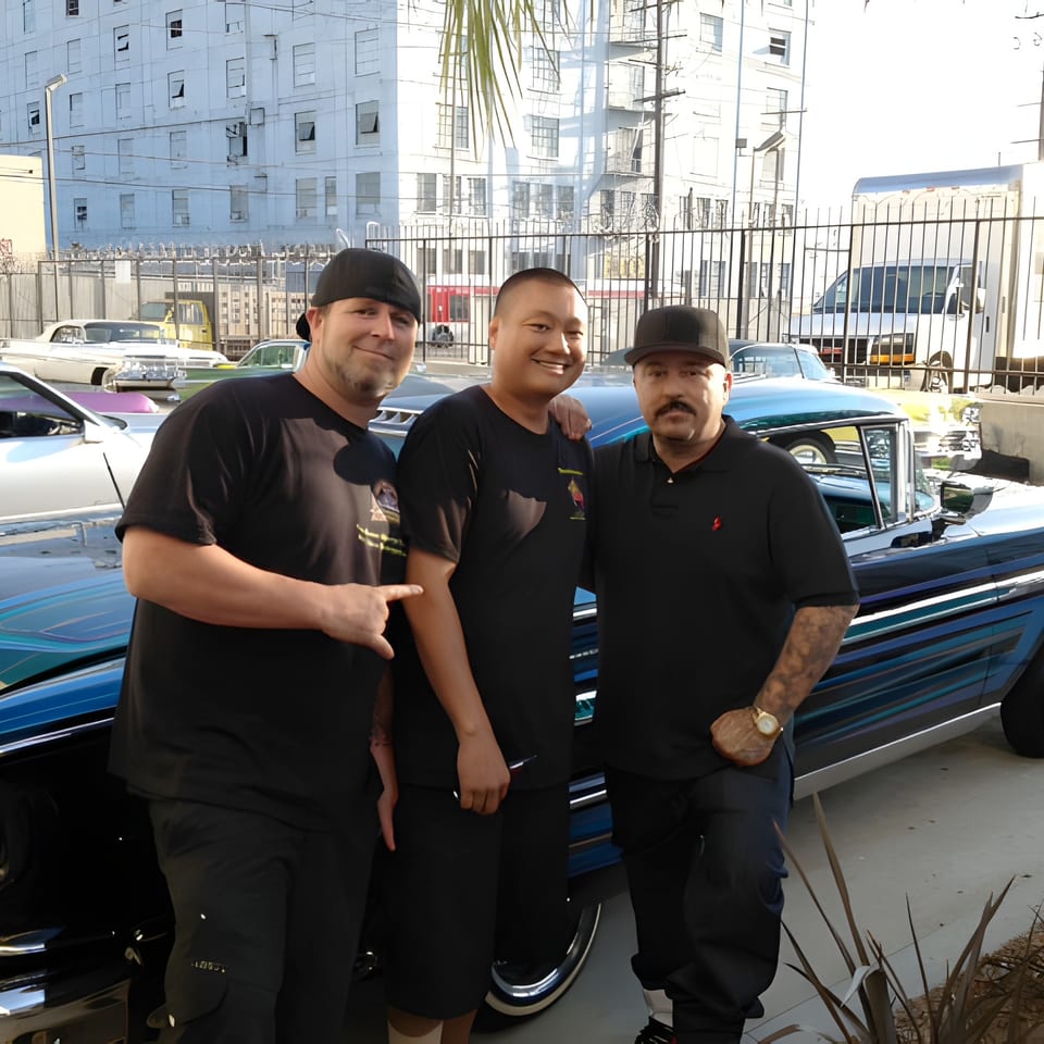 Three men smiling and posing next to a custom blue lowrider car in an urban setting.