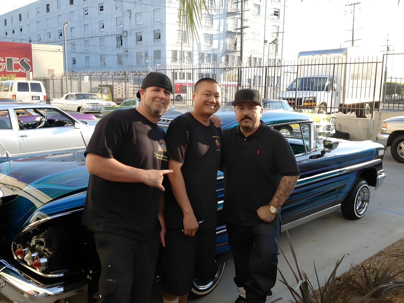 Three men smiling and posing next to a custom blue lowrider car in an urban setting.
