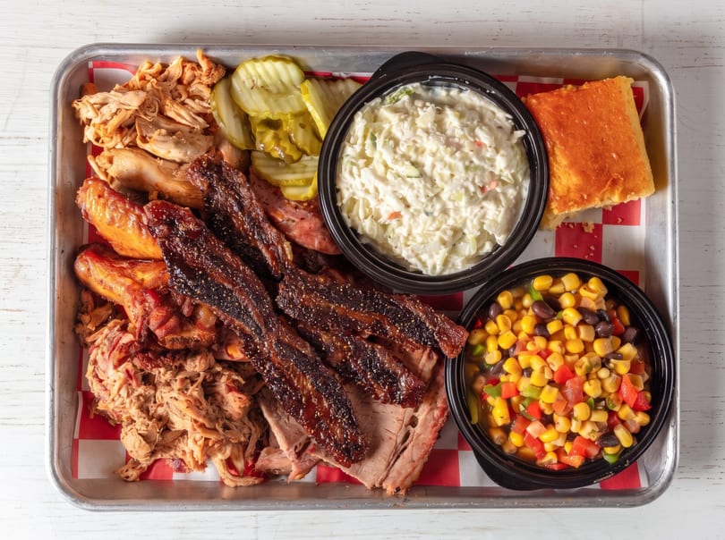 Large barbecue platter featuring pulled pork, brisket, chicken, coleslaw, cornbread, and corn salad.