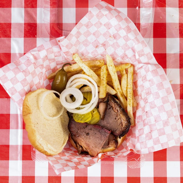 Open brisket sandwich with fries, pickles, onions, and a jalapeño on a red checkered table.