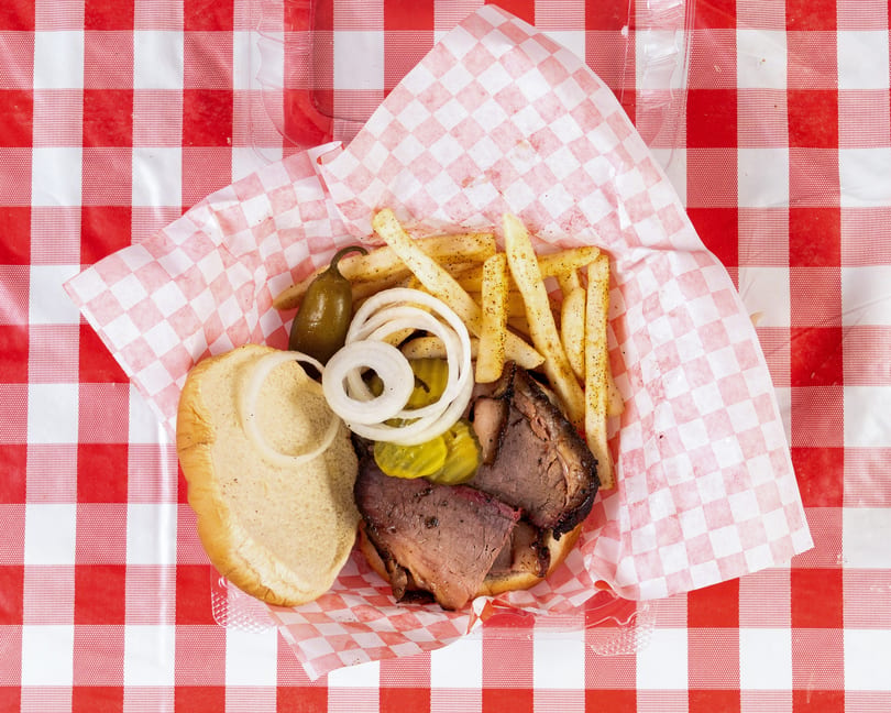 Open brisket sandwich with fries, pickles, onions, and a jalapeño on a red checkered table.