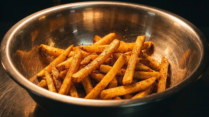 Close-up of golden-brown seasoned french fries in a metallic bowl, under warm lighting.