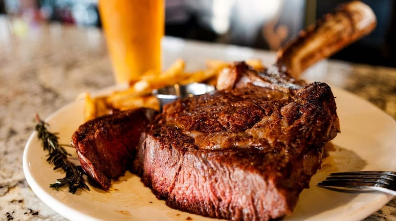 A perfectly cooked tomahawk steak with fries, a sprig of rosemary, and a drink on a marble counter.