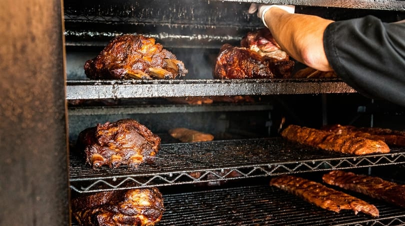 A gloved hand places ribs into a large, dark smoker filled with various cuts of meat.