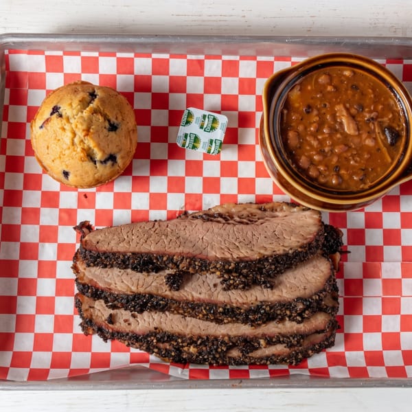 Smoked brisket slices, baked beans, and a blueberry muffin served on a checkered tray.