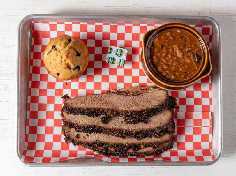Smoked brisket slices, baked beans, and a blueberry muffin served on a checkered tray.
